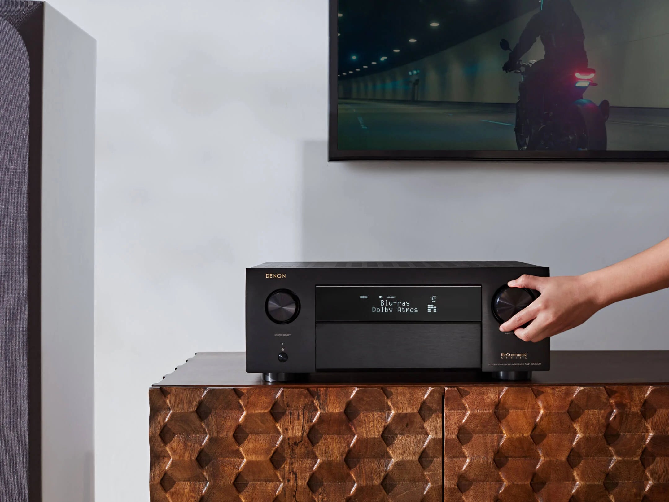 Audio receiver on a wooden cabinet with a television displaying a motorcycle scene in the background.