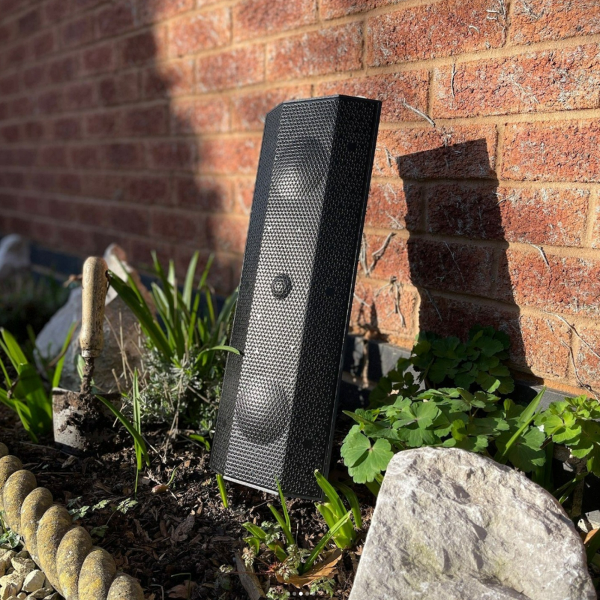 Black outdoor speaker against a brick wall with plants and stones.