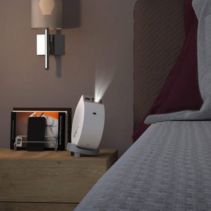 Small white projector on a nightstand next to a bed with a lamp in the background.